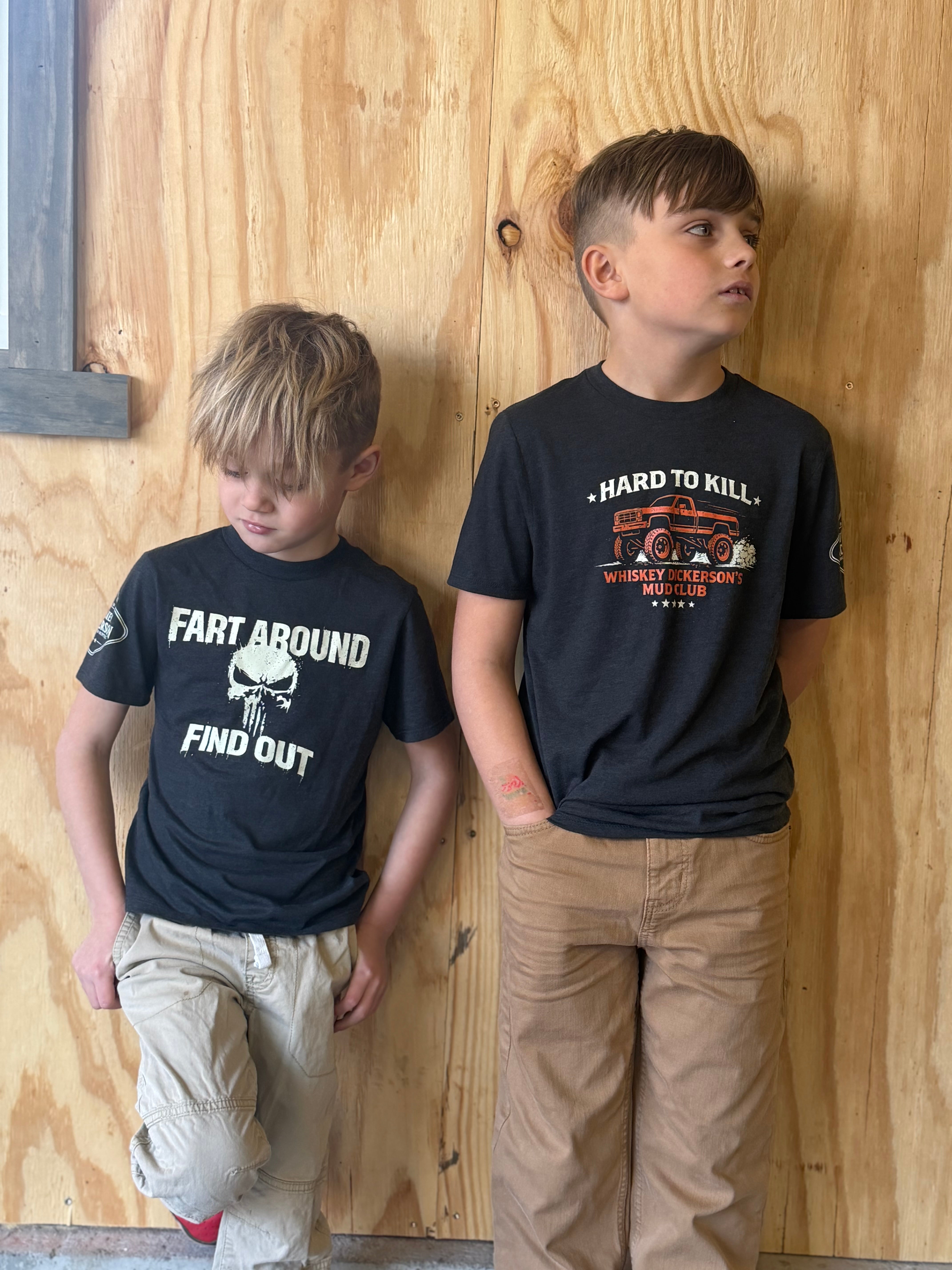 Two children wearing black Whiskey Dickerson t-shirts with humorous text designs against a wooden wall.